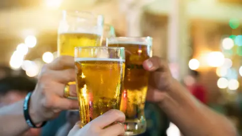 Several people are clinking large glasses of beer. The background is lit with soft, blurred lights.