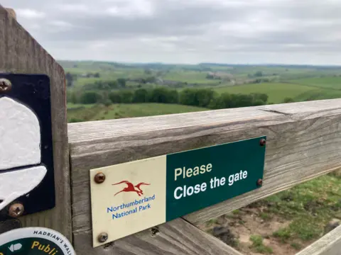 A cream and green "Northumberland National Park: Please close the gate" sign is nailed to a wooden gate. Lush, green fields stretch out behind it.