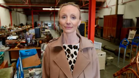Laurie Rossiter is wearing a brown trench coat and is standing in the auction hall, with all of the goods lined up behind her.