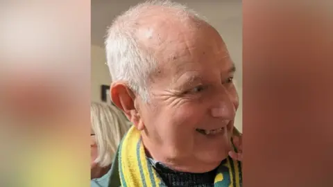 Jim Davies is pictured smiling. He is balding, with some short, white hair and is wearing a yellow and green football scarf.