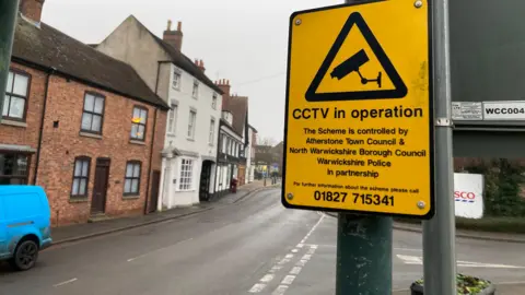 A yellow CCTV warning sign on a metal post in a street with a number of brick and white-painted houses on the opposite side of the road.