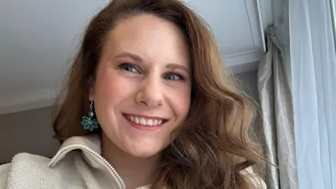 Contributor picture A selfie of Molly Leonard. She has long, brown wavy hair and drop earrings with a turquoise flower on. She is wearing a white fleece. 