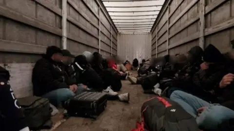About 20 people sat on the floor of the back of a lorry on wooden boards. Their faces are obscured either by blurring or jumper hoods, or caps.