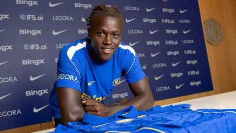 Quenda resting his arms on a table with a Chelsea shirt on and in front of him after signing for the club