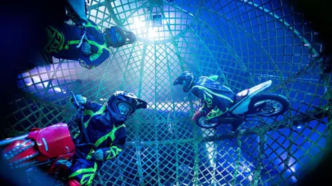 Ben Birchall/PA Media Three motorcyclists ride around inside a giant wheel as they rehearse for performances at Circus Vegas on the Downs in Bristol. They are wearing illuminated patches on their motorbike leathers and are lit up by spotlights