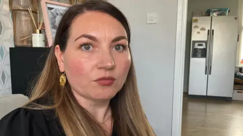 Liz Charlton, sitting on a sofa in her home, looking serious, during a conversation with BBC health correspondent Mark Norman. To her right, there is a doorway into her kitchen and a tall, silver fridge can be seen.