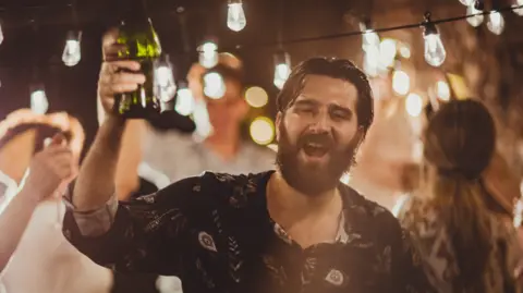 A bearded man in his 30s lifts a bottle of booze at a backyard party with fairylights