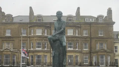 Jamie Niblock/BBC The statue of Benjamin Britten in front of a building divided into multiple properties. A flag pole with the Union Jack flag is also in the background.