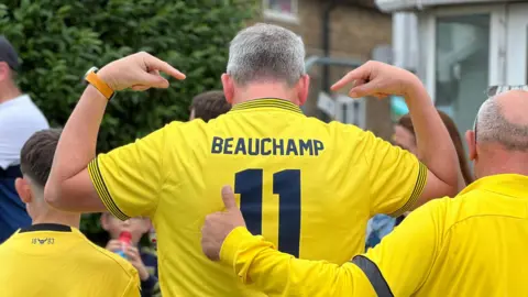 A man wearing a yellow Oxford shirt with Beauchamp 11 on the back. He is pointing at the name.