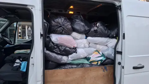The side of a large white van with the sliding door open, revealing a cargo area packed almost to the roof with numerous black and white plastic bags. The bags appear to contain soft items such as clothing, bedding, pillows, duvets, or other textiles.