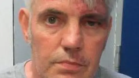 West Yorkshire Police David Hill, who has short, neatly trimmed grey hair and is wearing a light grey crew-neck shirt. The background consists of a wall with two distinct sections: one side is white, and the other is a darker blue panel. There is also a metallic door hinge or frame visible on the right side of the image.