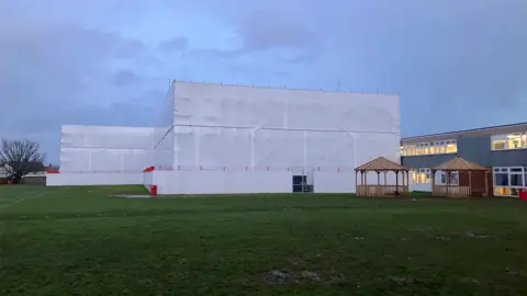 White sheeting covered scaffolding around a school building in a playing field at dusk with the lights on in some neighbouring classroom buildings and two wooden gazebos on the field