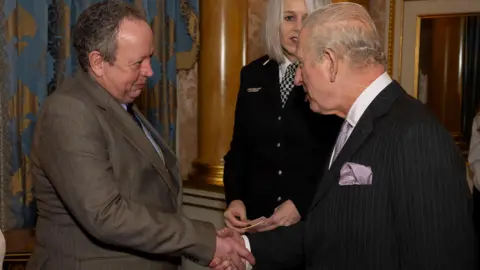 PA Media Train driver Andrew Johnson, wearing a brown blazer, smiles as he shakes hands with the King, King Charles, wearing a black suit,