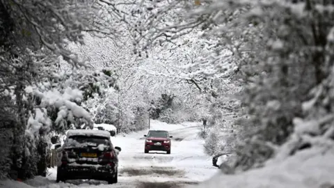 Getty Images A car drives down a snowy country road which is framed by trees. Another car is parked at the side of the road.