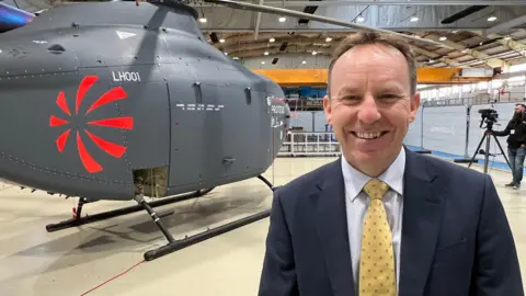 A man is wearing a navy blue suit jacket, a blue shirt and a yellow tie, and smiling at the camera. Behind his right shoulder is a helicopter. They are in a hangar.