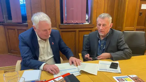 BBC Council leader George Duggins and finance cabinet member Richard Brown sit at a desk in a wood-panelled room, looking over tables of figures and other paperwork. Both men are wearing suit jackets with open-necked shirts. 