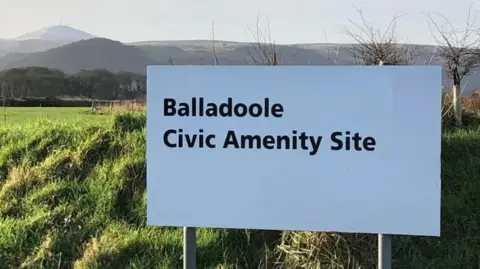 A large white sign with the word Balladoole Civic Amenity Site written on it in black lettering, in a grassed area with green hills in the background.