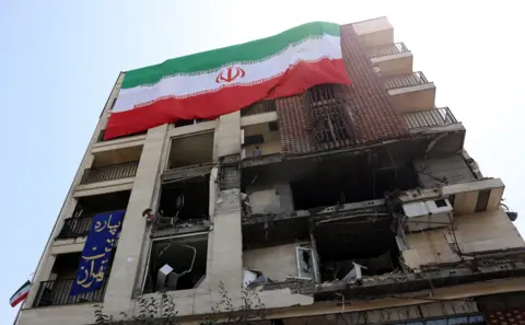 Abedin Taherkenareh/EPA/Shutterstock Uma bandeira nacional em larga escala está pendurada em um prédio danificado que foi atingido por um recente ataque aéreo israelense, em Teerã, Irã.
