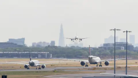 Two planes driving away from the camera on a runway at Heathrow Airport, with buildings including the Shard in the background. Another plane is facing the camera and coming in to land