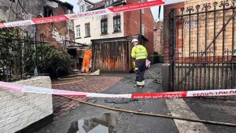 Fire engines and firefighters at the scene of a substation fire in Exeter. There is red and white tape creating a cordon, and two men wearing high-vis clothing.