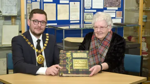 A woman - Helen Morrison - with a tartan scarf and white hair, holding a a book with a tartan cover and titled Greetings to the women of Leningrad on the front. Sitting alongside her is a man in a suit with short dark hair, glasses and a lord provost's chain.
