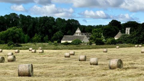 MONDAY - Round hay bales are in the foreground in a field of freshly cut grass. Behind you can see three grey stone buildings nestled in dark green trees. There is a blue sky overhead with white clouds.