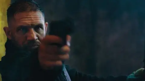 Netflix Tom Hardy in character, pointing a gun at the camera. He has short dark hair and is wearing all black.