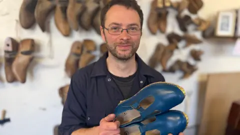 A man in a navy boiler suit with short dark hair and glasses holds up a pair of blue clogs.