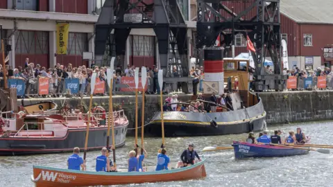 Bristol Harbour Festival Four boats on the water in Bristol. There is a large crowd standing at the harbour edge watching them sail.