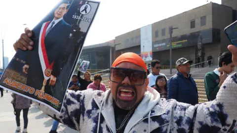 Getty Images A supporter of Alberto Fujimori wearing an orange headgear, sunglasses and a blue and white patterend jacket expresses his grief while holding an image of the former president.