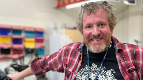 Robert Bierton is standing on the right. He has short dark blonde hair and is wearing a black T-shirt with a graphic on it, a red tartan shirt and a blue lanyard. He is leaning on a bike which is on the left.