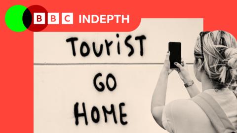 A sign that reads "tourist go home" and a woman photographing it