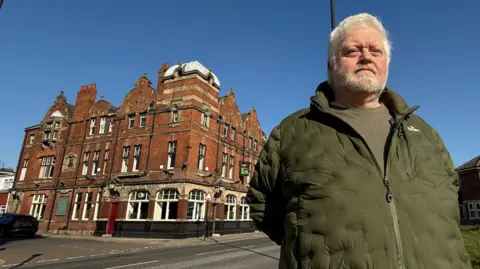 A man with white hair and a beard, wearing a khaki green puffer jacket and a green T-shirt, stands to the right of a red-brick period building.