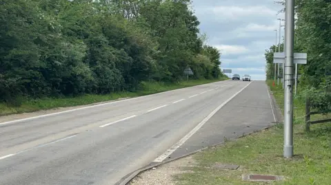 Two cars are travelling neck-and-neck along a dual carriageway towards the camera. The road is bordered by greenery on both sides and there are lamp posts on the right-hand grassy verge 