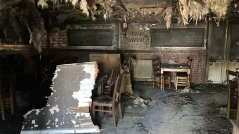 Remnats of the restaurant after the fire. The back wall display board is charred, with the words 'The Shed' still visible. The remaining furniture is surrounded by ash and debris.