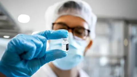 Getty Images Naukowcy noszący niebieskie rękawiczki, płaszcz laboratoryjny i sieć włosów zawiera fiolkę zawierającą przezroczystą płyn