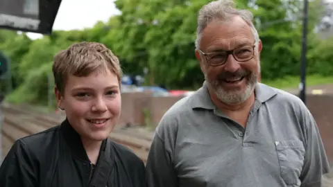 Henry and Dave Victor smiling. Henry is on the left and has light brown, short hair and is wearing a black jacket and smiling into the camera. Dave is on the right and has grey, short hair which is sticking up slightly at the top and is wearing a grey polo shirt and dark rectangular glasses. He is smiling while looking to the right. They are standing on a train platform with brown tracks visible below. There are trees out of focus in the background.