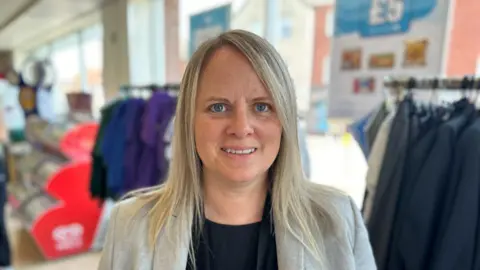 Helen Crouch in grey jacket with uniforms on racks in the background