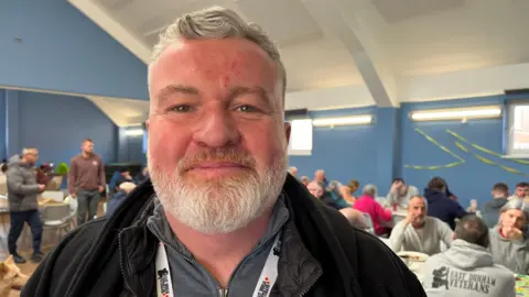 Lee Martin stands wearing a black fleece and a grey zip up top in front of a blue wall. He has grey hair and a beard. In the background people are sitting at tables in a hall with blue walls.