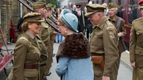 Keighley and Worth Valley Railway People in vintage World War Two military uniforms standing on a train platform, forming a line. A woman in a blue coat and hat with a fur scarf speaks to one soldiers. There is a steam train behind.