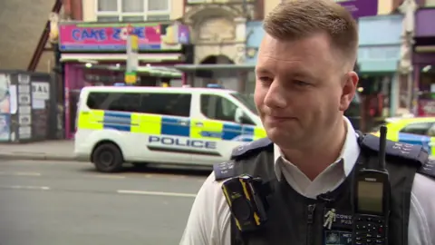 PC Joe West stands in uniform across the street from a Met Police van