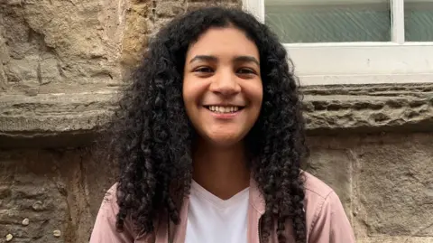 Chloe Aslett/BBC A woman with curly black hair, past her shoulders. She is smiling at the camera. She has a white t-shirt on and pink shirt-jacket over it.