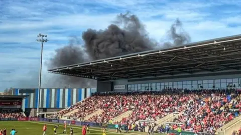 Charlie Putt The image shows a football match taking place inside a stadium, with players spread across the pitch and a large crowd filling the stands. Above the roofline at the far side of the ground, thick black smoke can be seen rising into the sky. A section of the stadium roof and structural beams frames the top of the image.