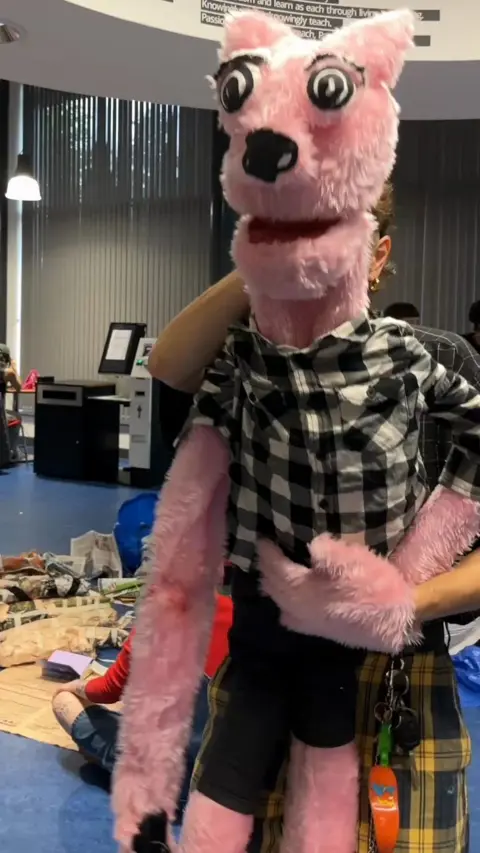 A large pink puppet in a black and white checked shirt, being held in front of a person