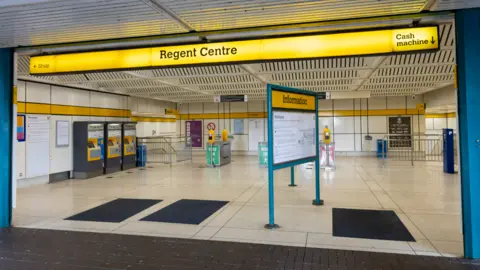 Regent Centre Metro station has a wide open concourse with a white and black marble floor and three ticket machines to one side. Open gates lead to the platform. 