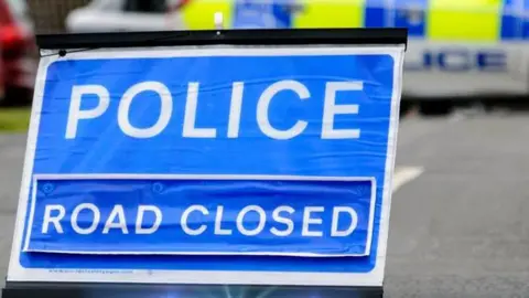 A blue road closed sign, with Police written on it, with a blurred police car behind it, on a road. 