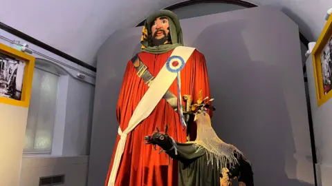 BBC A figure a few meters high, bearded male face with a hat and wearing a long red coat and white sash, next to a smaller dark coloured horse-type figure