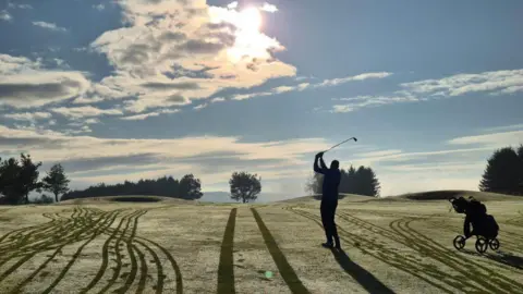 Ben Walls A golfer is silhouetted on a course on a sunny morning, with buggy tracks visible on the fairway.