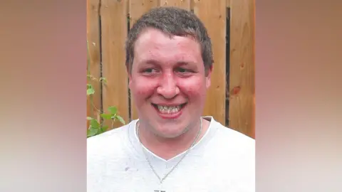 Steven Peck. He has short light brown hair, wearing a white t-shirt and silver chain with a cross.