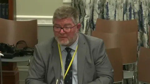 A man with grey hair and glasses sitting at a desk. He is wearing a grey suit with a navy tie and a yellow lanyard.  